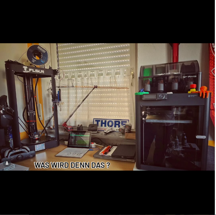 Mehrere 3D-Drucker stehen in einem Arbeitsbereich, dar&uuml;ber der Schriftzug „Was wird denn das?“. 